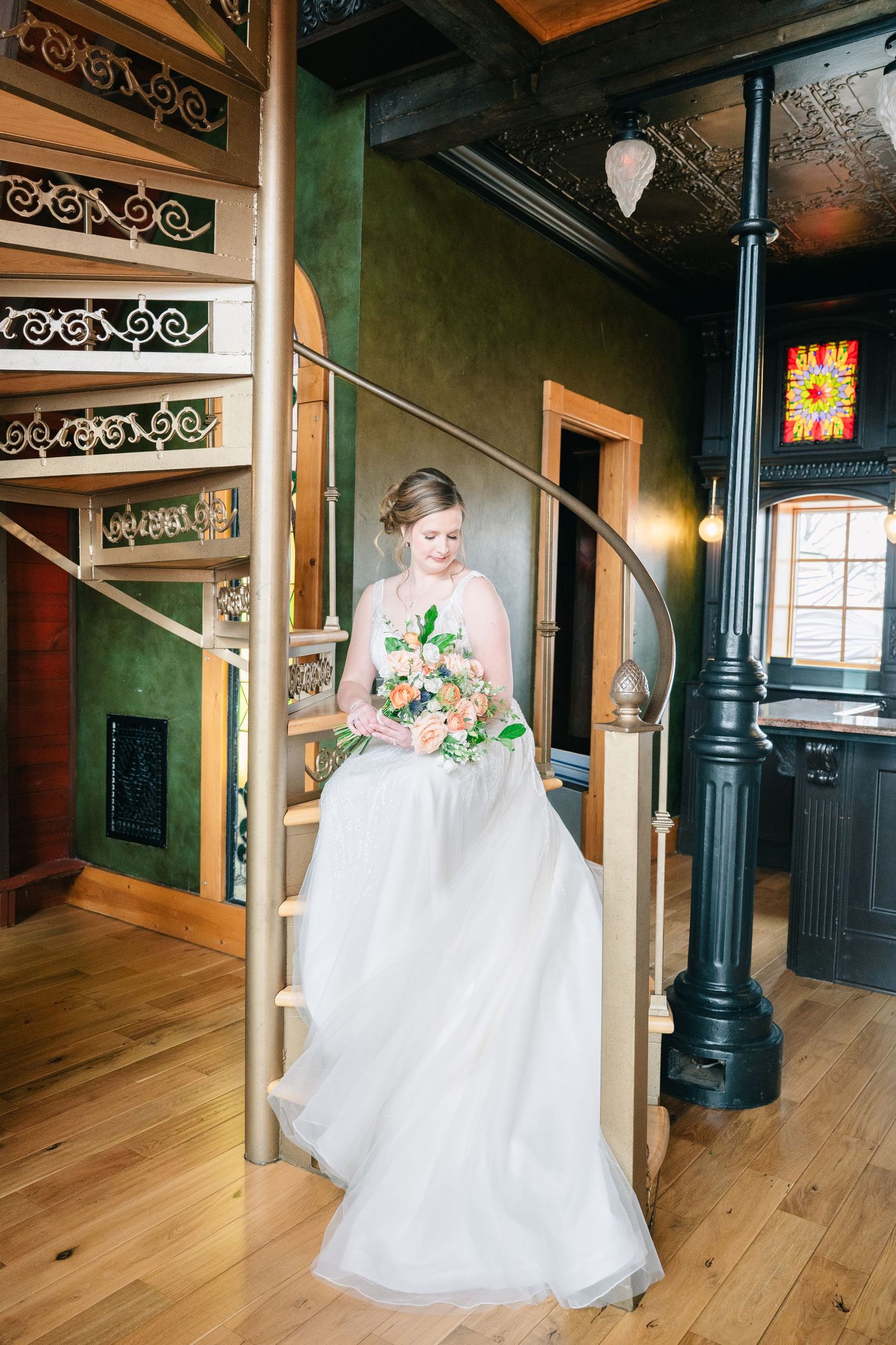 Bride in white gown holding peach bouquet descending ornate spiral staircase in elegant venue.