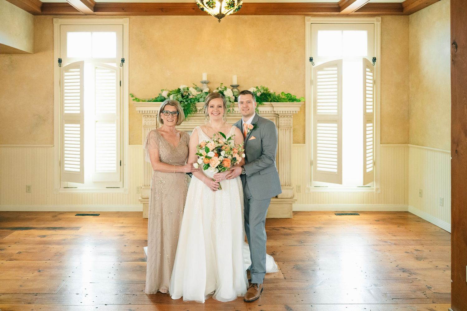 Bride in white gown holding peach bouquet posing with two family members in front of fireplace mantel.