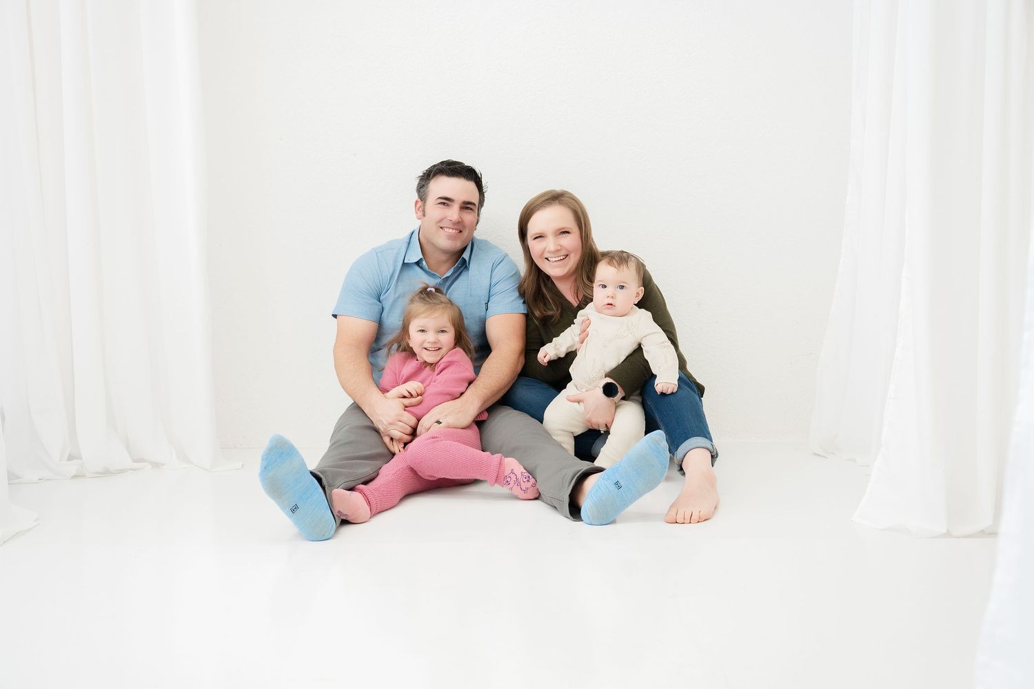 Happy family of four sitting on white floor, parents smiling with toddler girl and baby in bright studio photo.