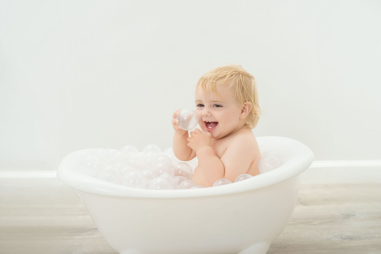 Happy blonde toddler playing with bubbles during bath time in a white bathtub.. Minnesota milestone photographer