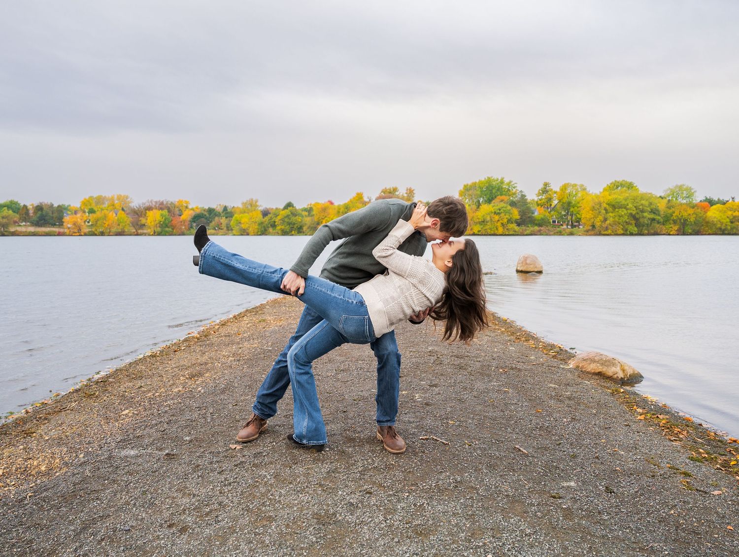 A couple embraces playfully along a scenic waterfront path during autumn with colorful fall foliage in the background at Lake Como. E sesh.