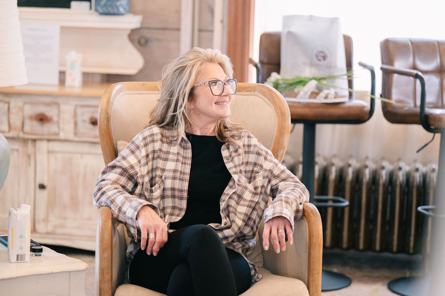Woman with gray hair and glasses relaxes in a beige armchair in a cozy, well-lit interior space.