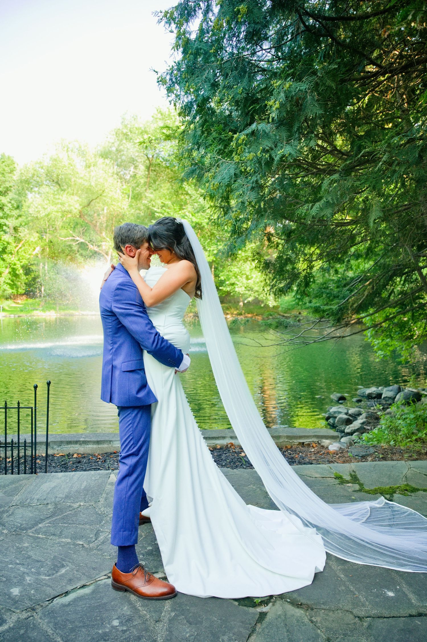 Bride and groom embrace near a serene pond surrounded by lush green trees on their wedding day.