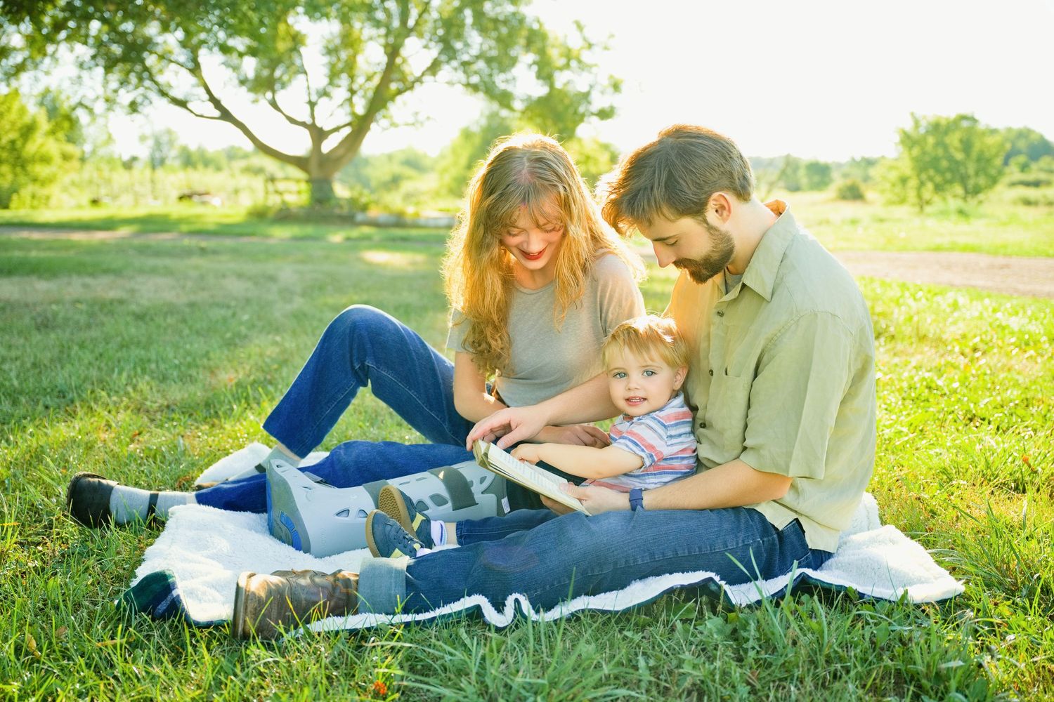 Family of three sitting on blanket in sunny park, parents reading book with toddler on lap outdoors.