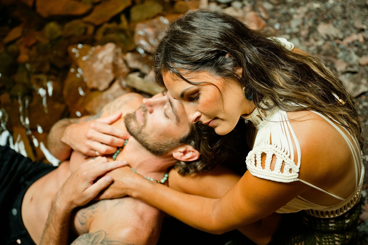 A woman in a white top leans over a man lying on rocky ground in a romantic outdoor embrace.