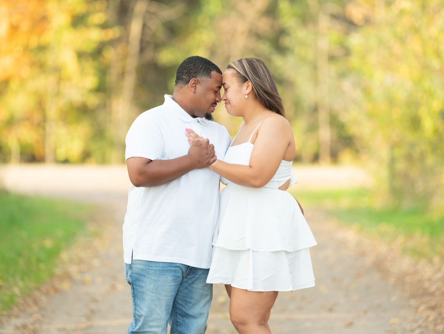 Central Minnesota Best Engagement Photographer - Couple in white outfits sharing a romantic moment on a tree-lined path with autumn foliage.