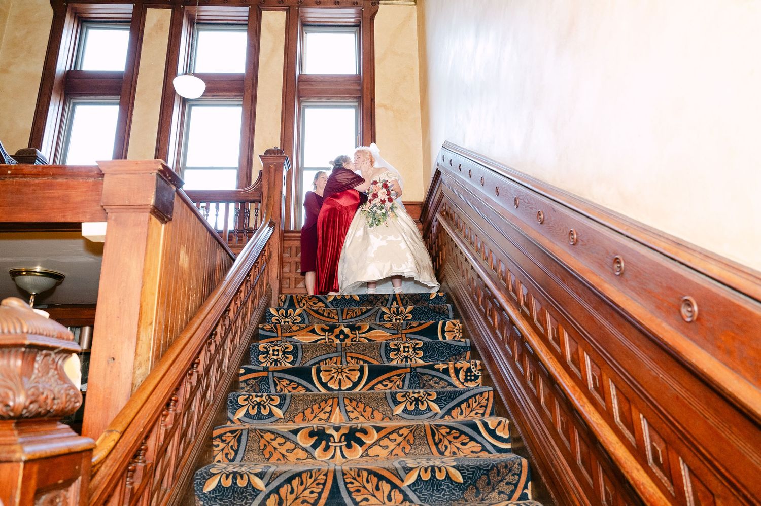 Central MN Wedding Photos | A bride in a white gown descends an elegant wooden staircase with ornate blue patterned carpet runner.