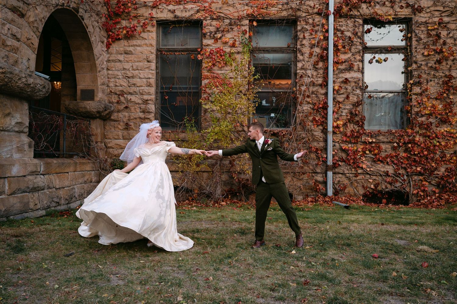 Central MN Wedding Photos | Bride and groom playfully dancing on lawn beside historic stone building.