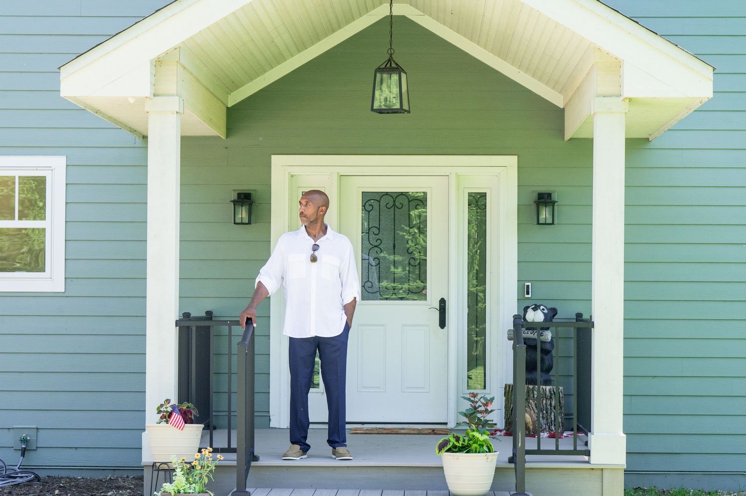 A man in a white shirt stands on the porch of a green house with white trim and a white front door.