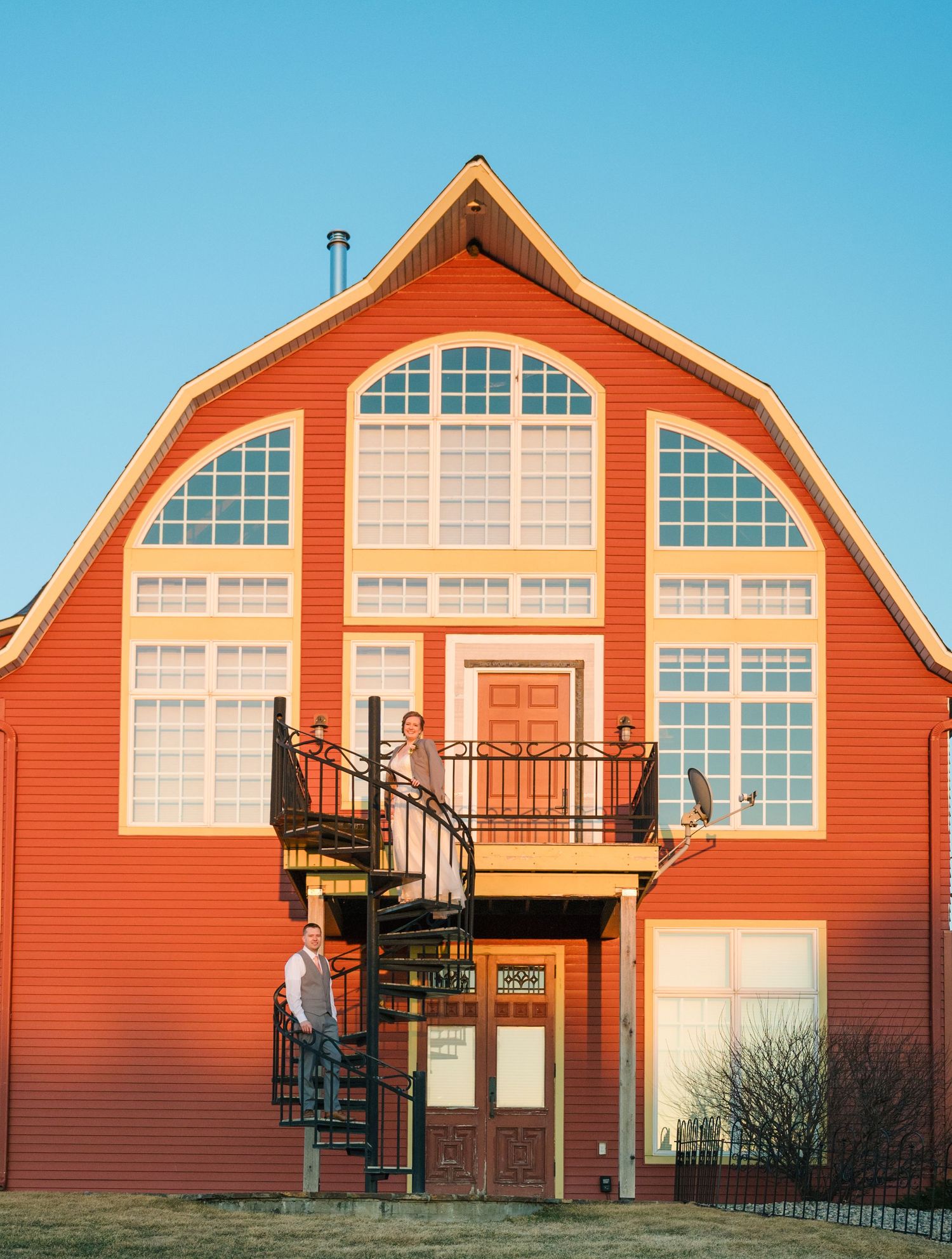 Couple stands on spiral staircase of large red barn-style wedding venue under clear blue sky.