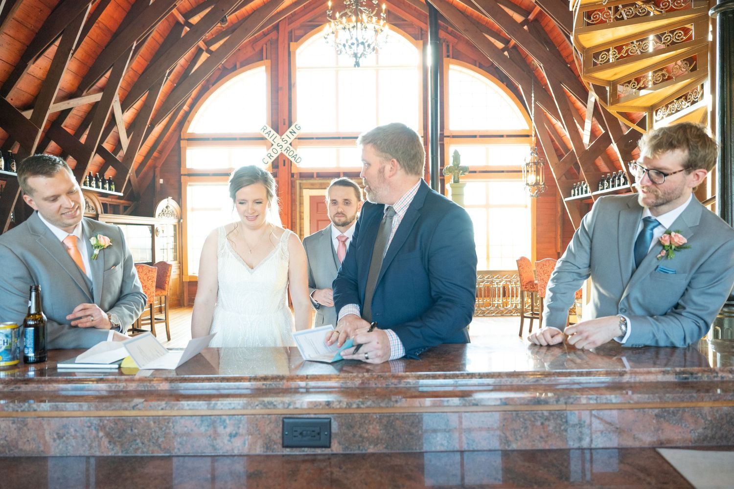 Bride and groom signing wedding documents at wooden bar with officiant and witnesses present.