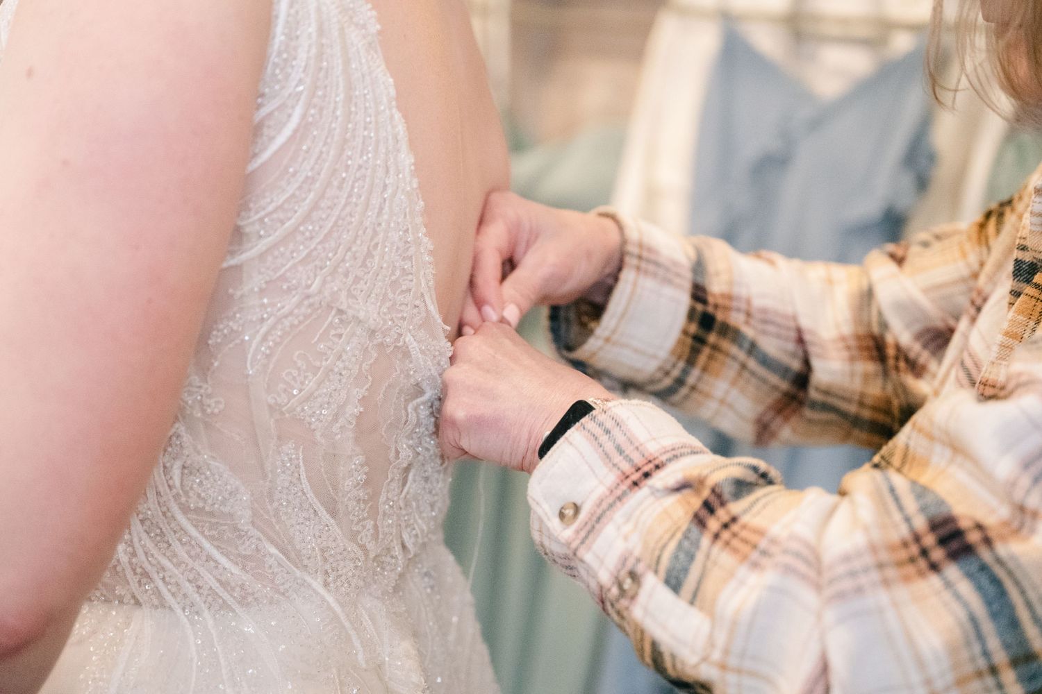 Helper fastening buttons on back of white lace wedding dress during bridal preparation.
