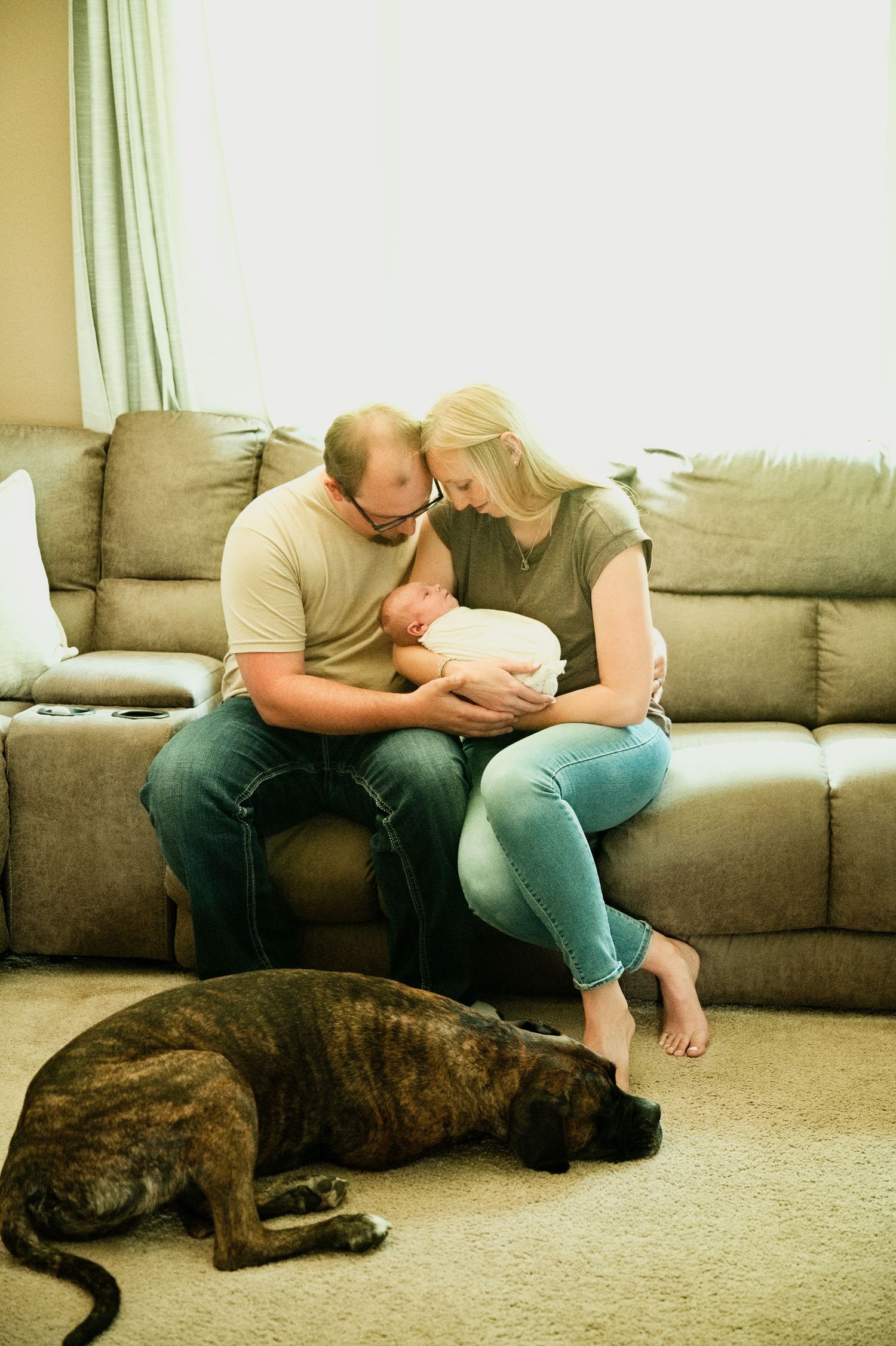 New parents sit on couch holding newborn baby while brindle dog rests on floor nearby.