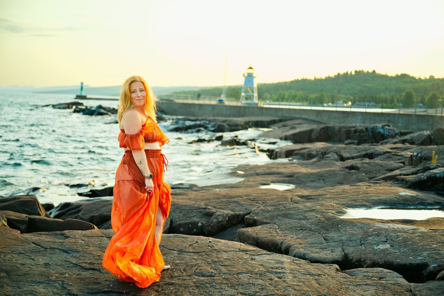 Woman with long blonde hair in a flowing orange dress poses on lakeside rocks with a lighthouse in the background.