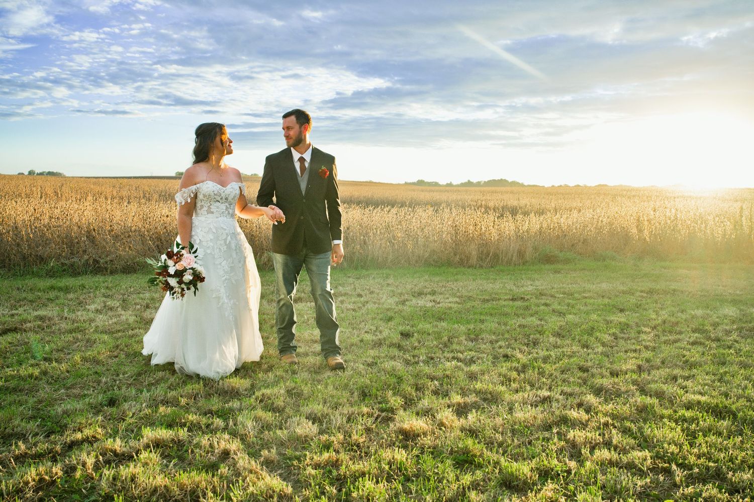 Bride and groom holding hands in a sunlit field of golden crops, bride in white lace gown holding bouquet.