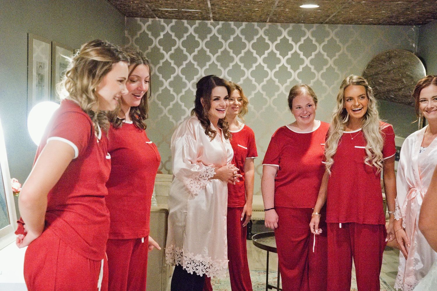 Bride in pink robe laughs with bridesmaids dressed in matching red pajamas during wedding morning preparations.