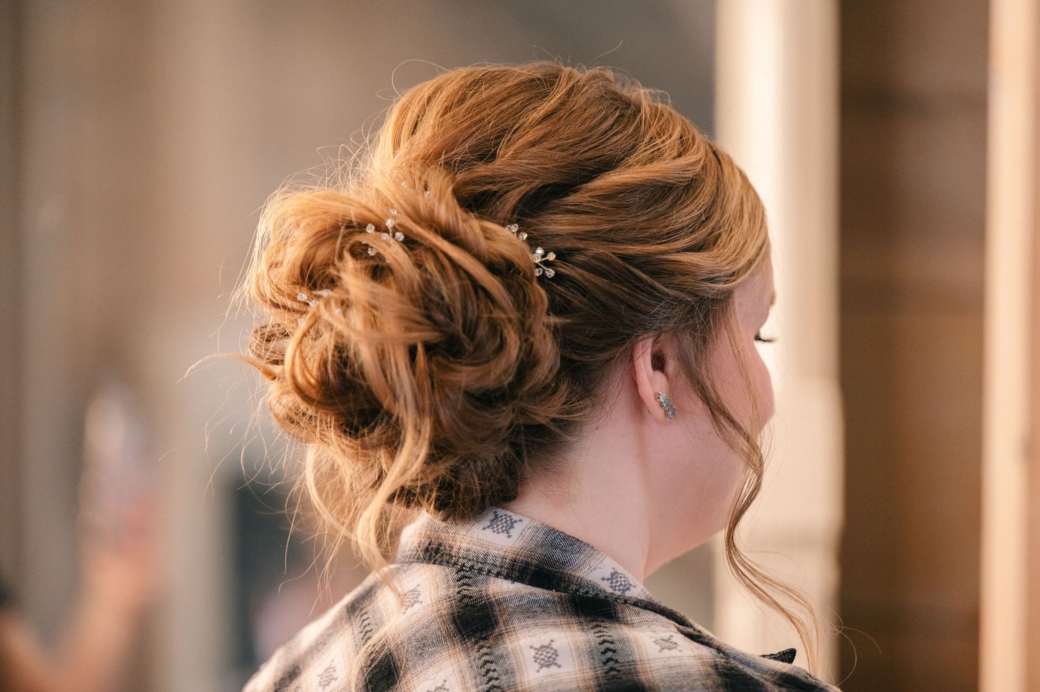Woman with elegant auburn updo hairstyle adorned with pearl pins, wearing a plaid shirt, photographed from behind.