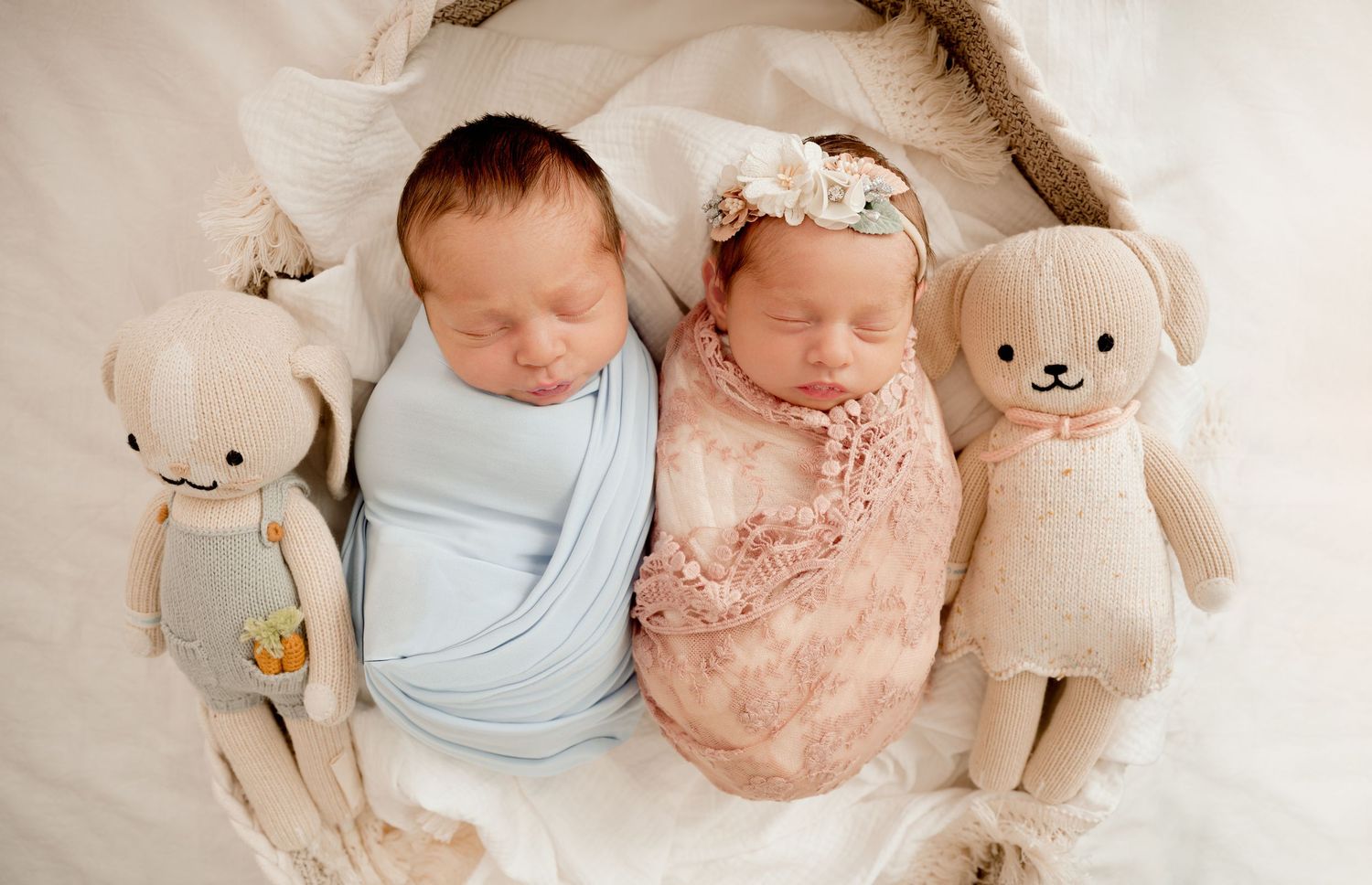 Two newborn twins swaddled in blue and pink wraps, nestled between stuffed teddy bears.