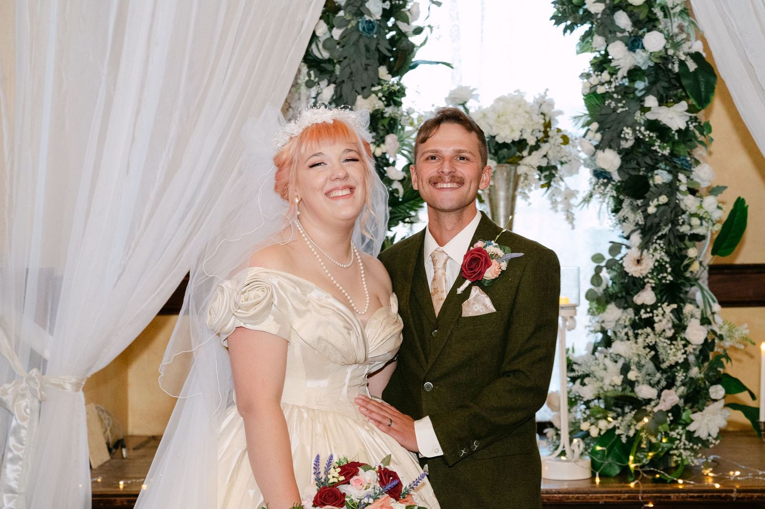 Central MN Wedding Photos | Smiling newlyweds pose at the altar decorated with white flowers and greenery after their wedding ceremony.