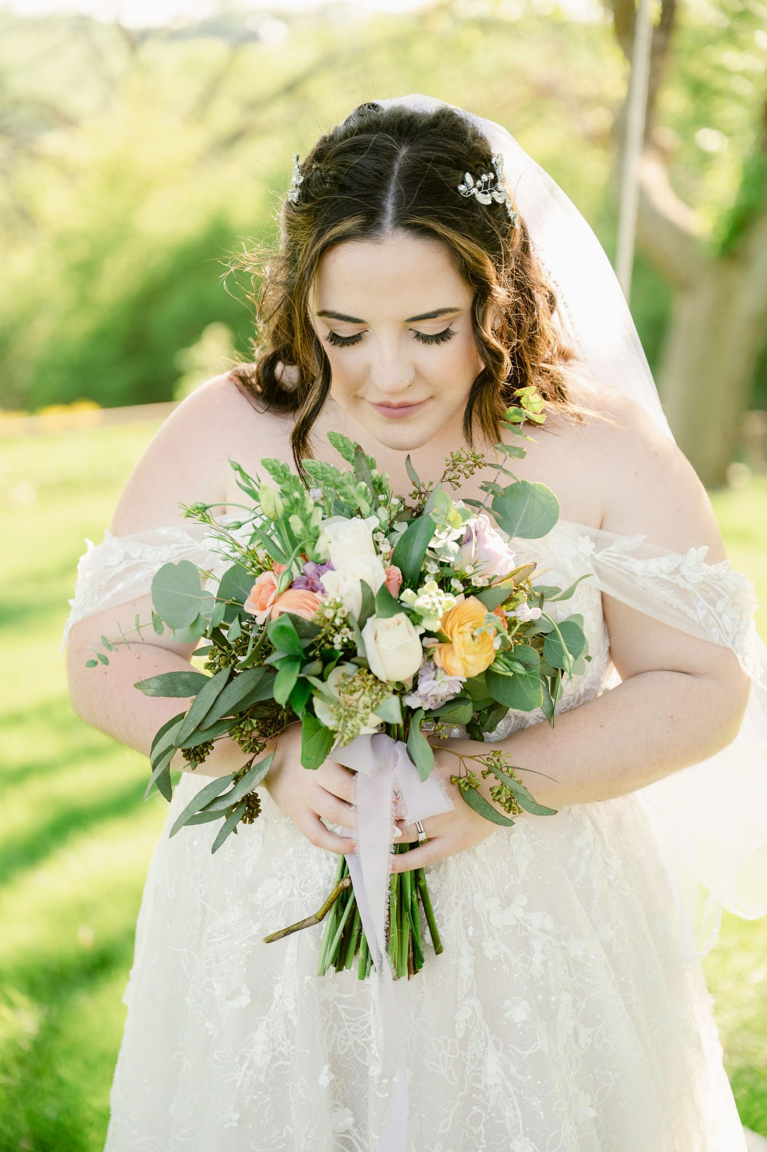 Central MN Wedding Photos - Bride in lace off-shoulder gown gazes at colorful bouquet of roses and greenery in sunny garden setting.