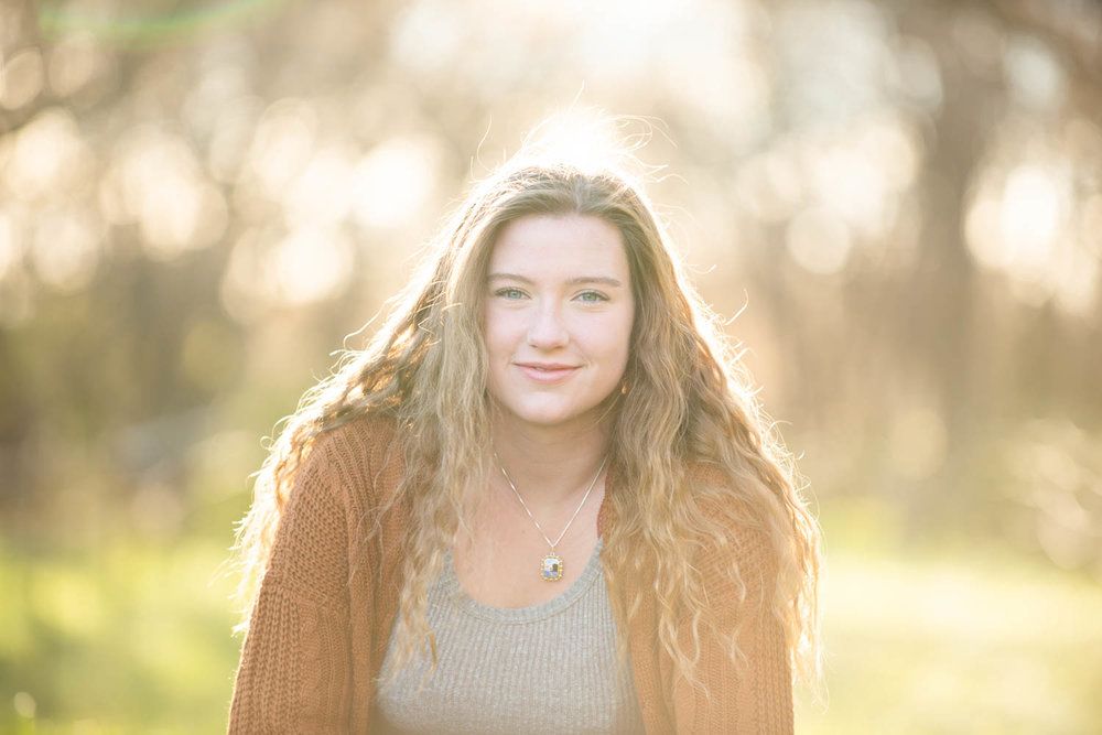 Central MN Senior Photography - Young woman with long curly hair wearing an orange cardigan smiling outdoors in soft golden sunlight.