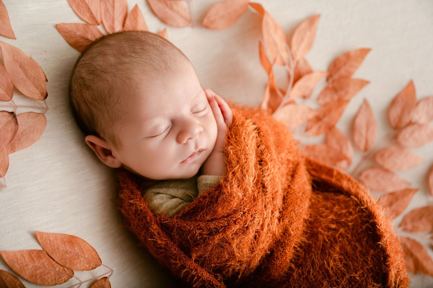 Baby sleeping on white blanket surrounded by orange autumn leaves and wearing knit sweater.