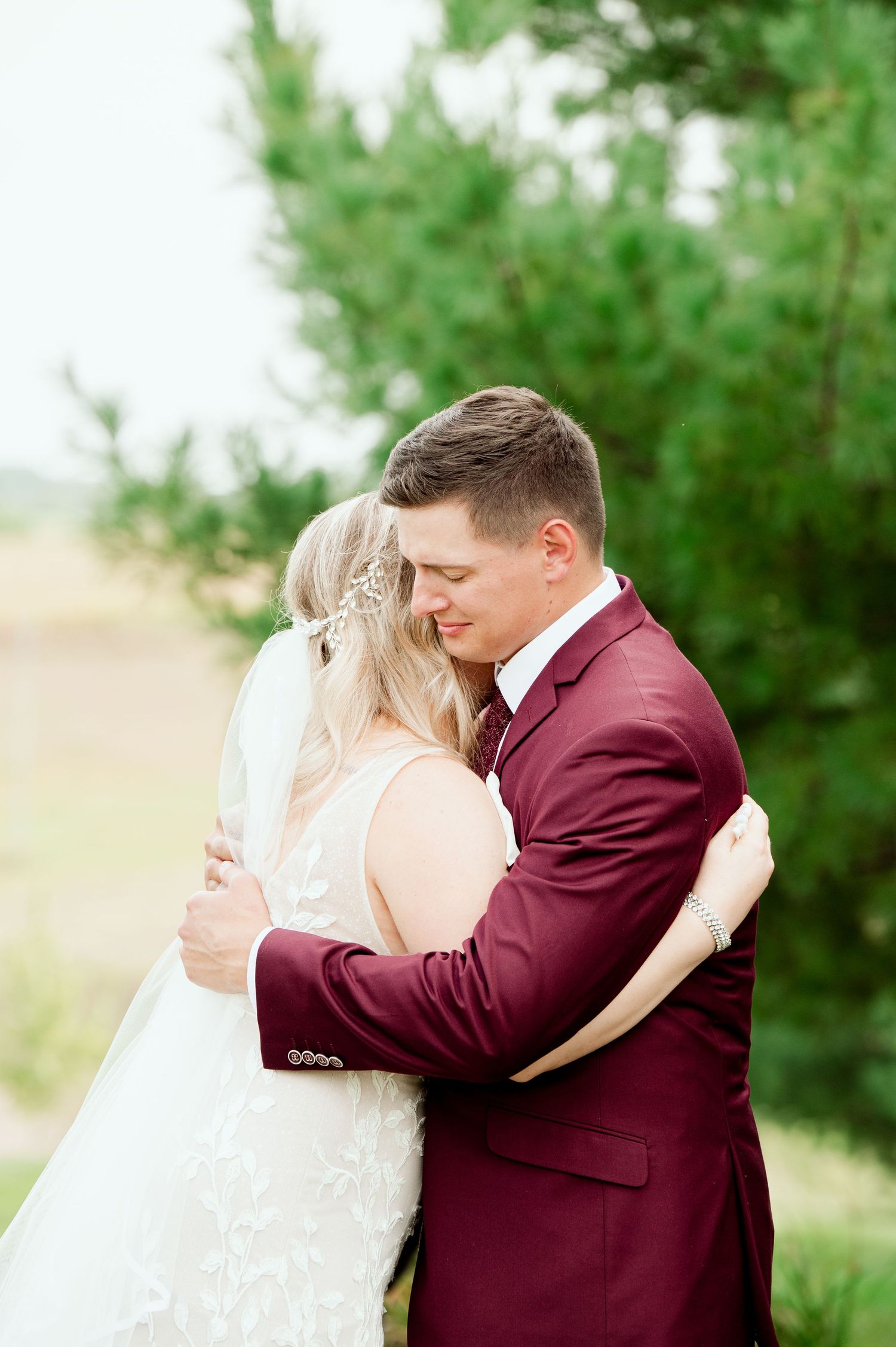 A romantic wedding couple embraces outdoors with the groom wearing a burgundy suit and the bride in white with a flowing veil.
