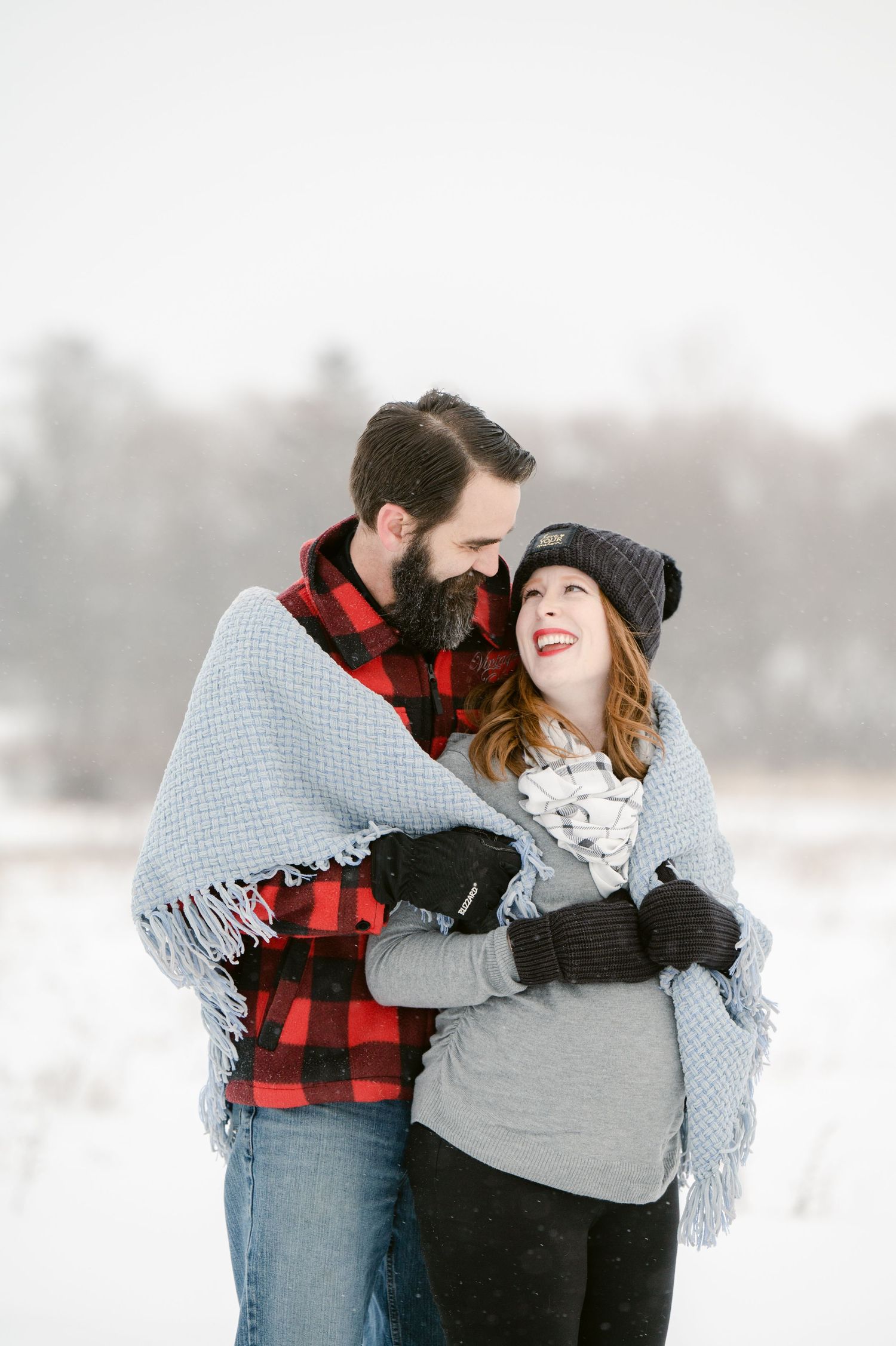 The Clark’s romantic winter maternity photoshoot series in Foreston playfully in a snowy landscape wearing buffalo plaid and a gray blanket.