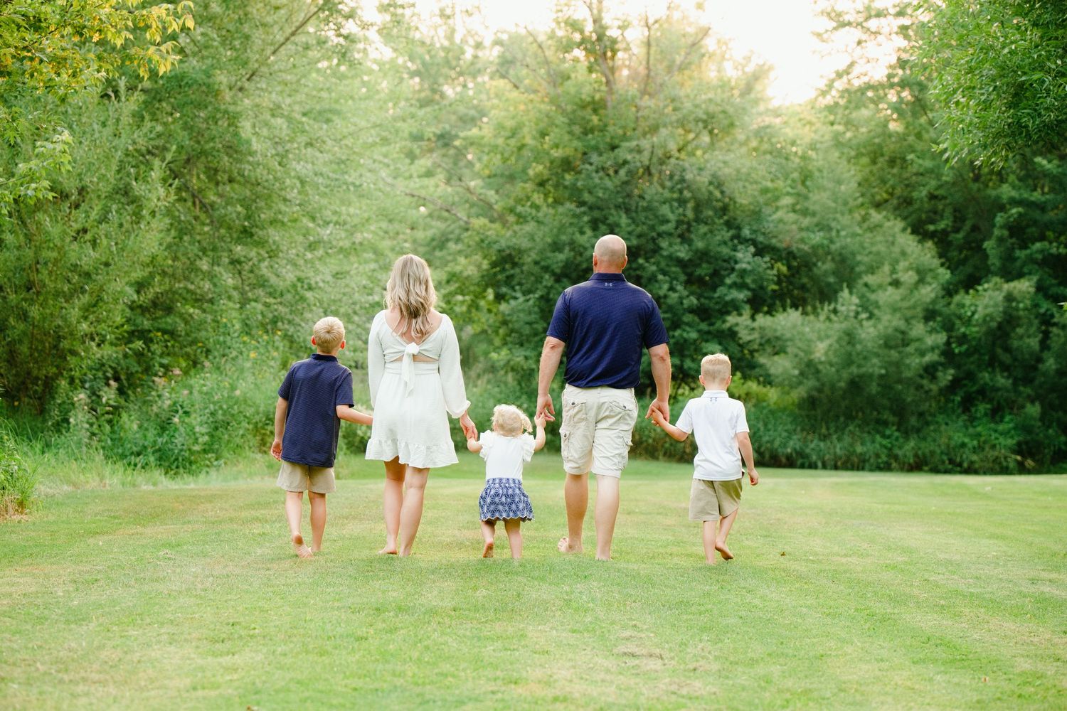 Family of five walking hand-in-hand across lush green lawn toward trees, viewed from behind on sunny day.