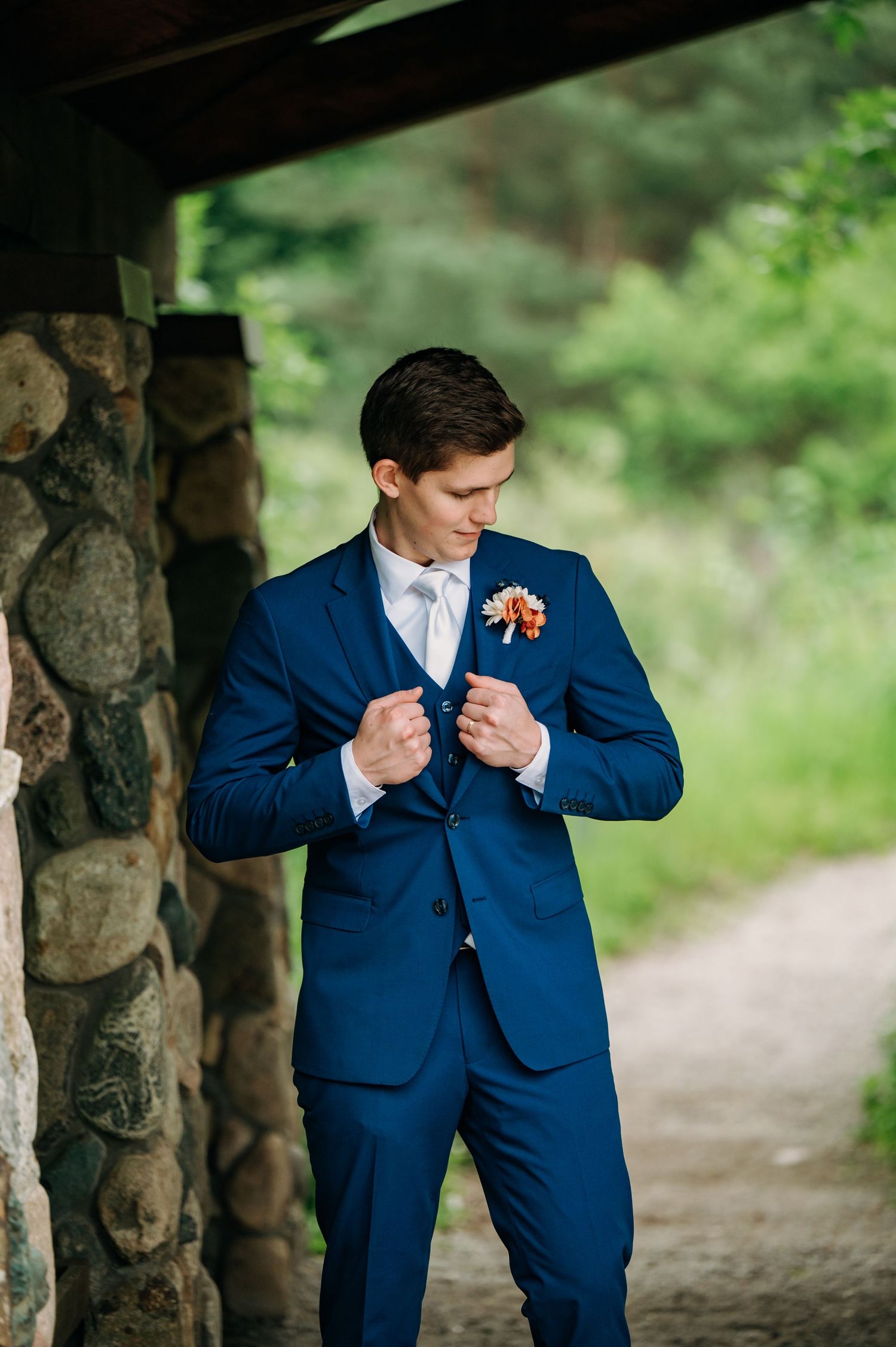 A groom adjusts his navy blue suit jacket while standing near a stone pillar outdoors.