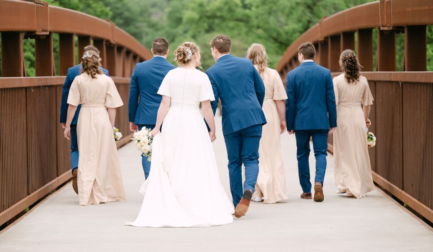 Wedding party walks together across a wooden bridge surrounded by lush greenery in matching blue and pink attire.
