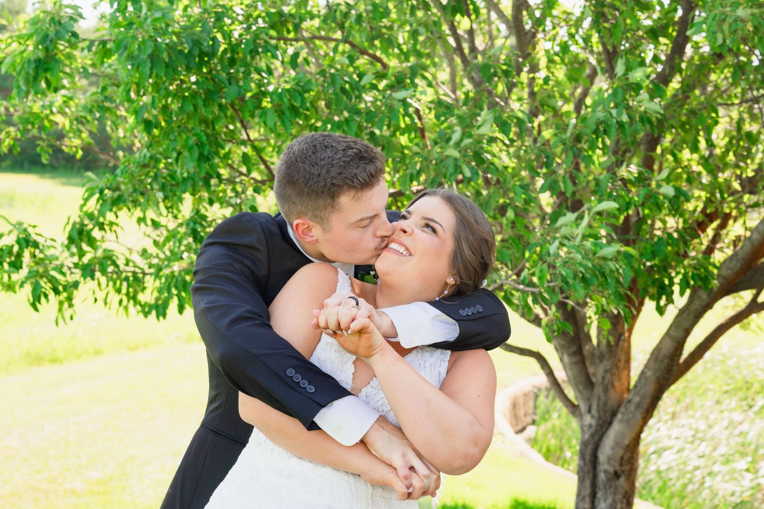 Groom in black suit lifts and embraces smiling bride in lace gown under a green tree.