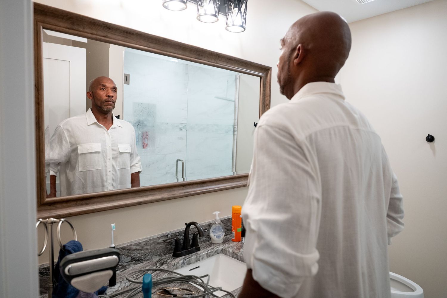 Man in white shirt examining his reflection in a bathroom mirror above a granite countertop.