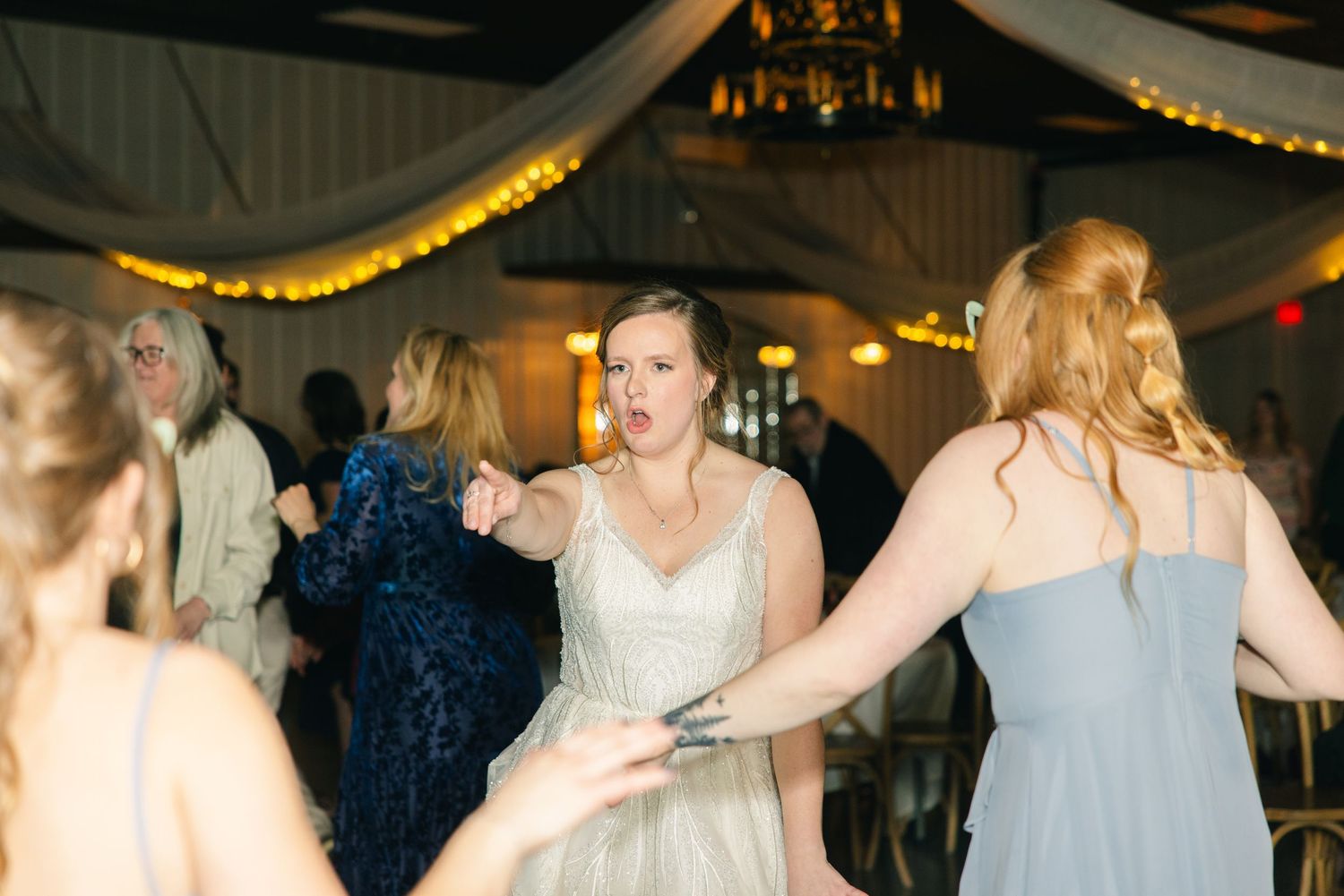 Bride dancing energetically with bridesmaids in sage green dresses at wedding reception.