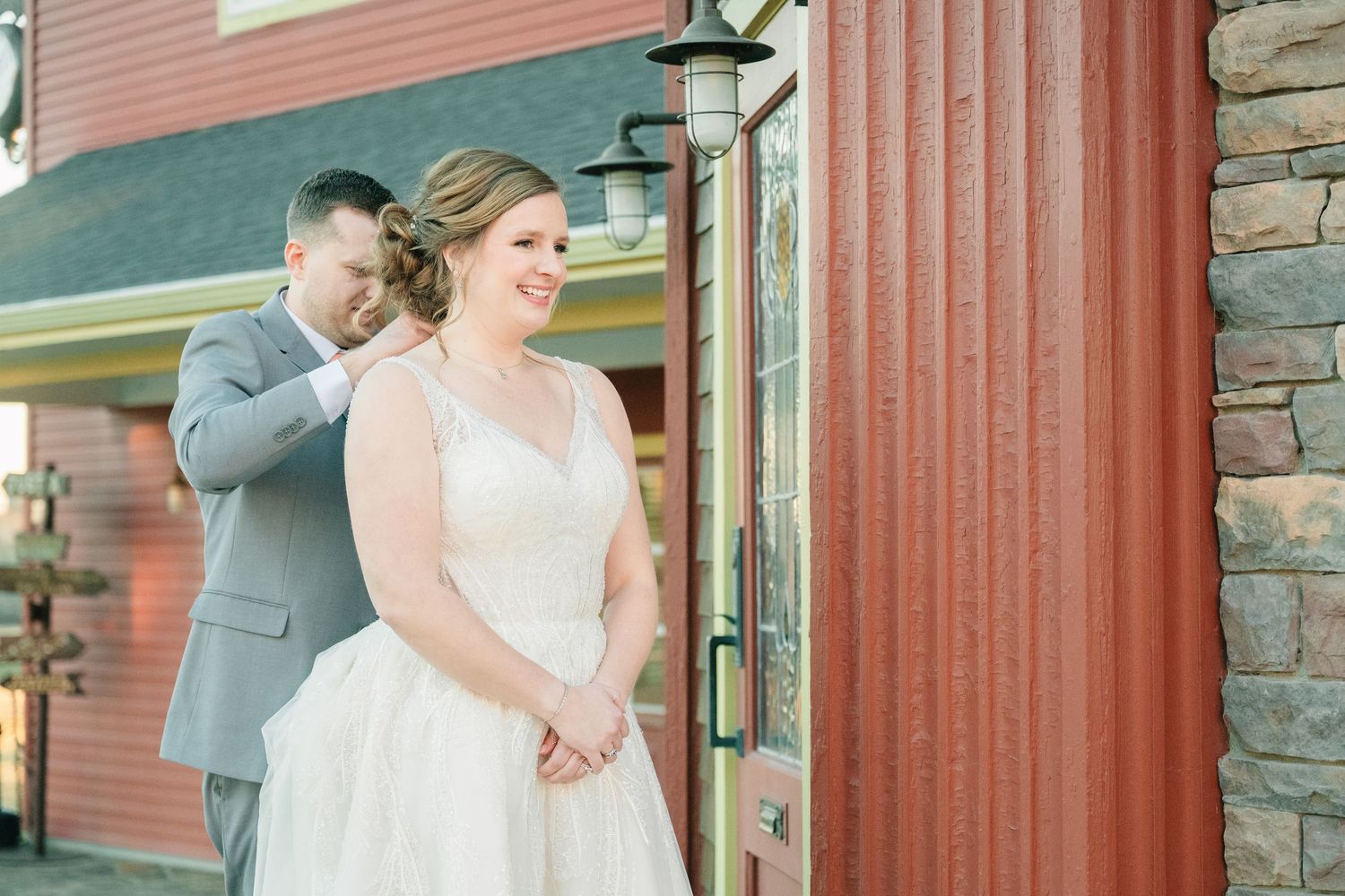 Groom tenderly embraces bride from behind during romantic first look outside rustic red barn venue.