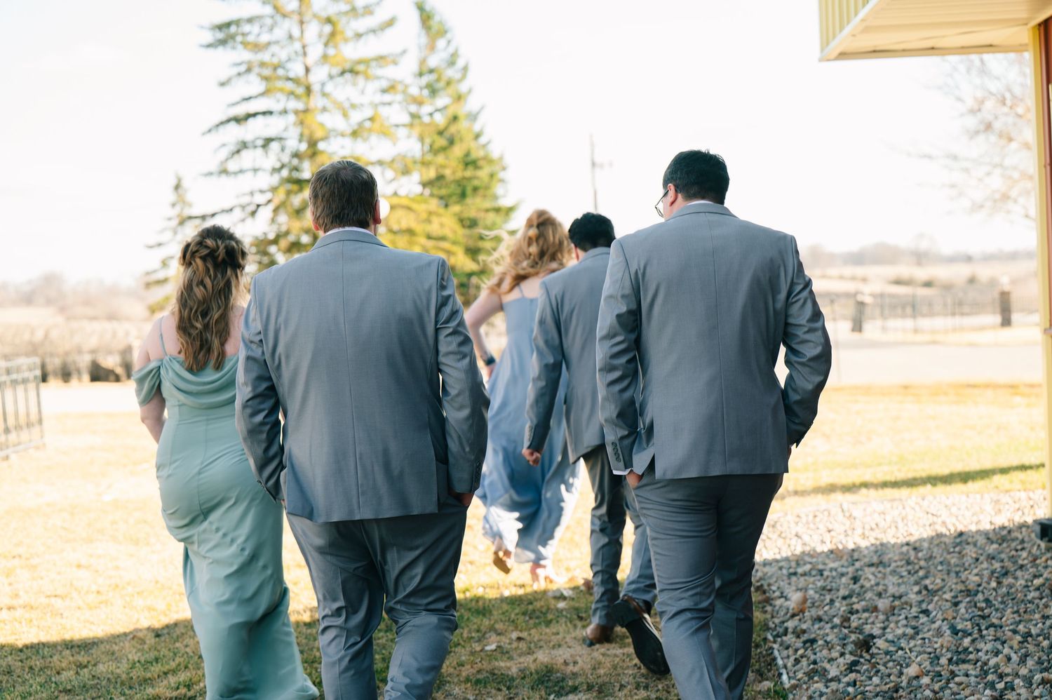 Wedding party walking together outdoors in gray suits and sage green dresses toward a building.