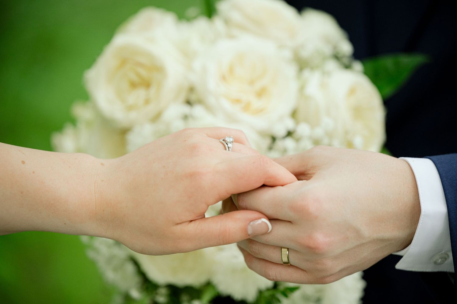 Close-up of hands exchanging wedding rings with white roses in background.