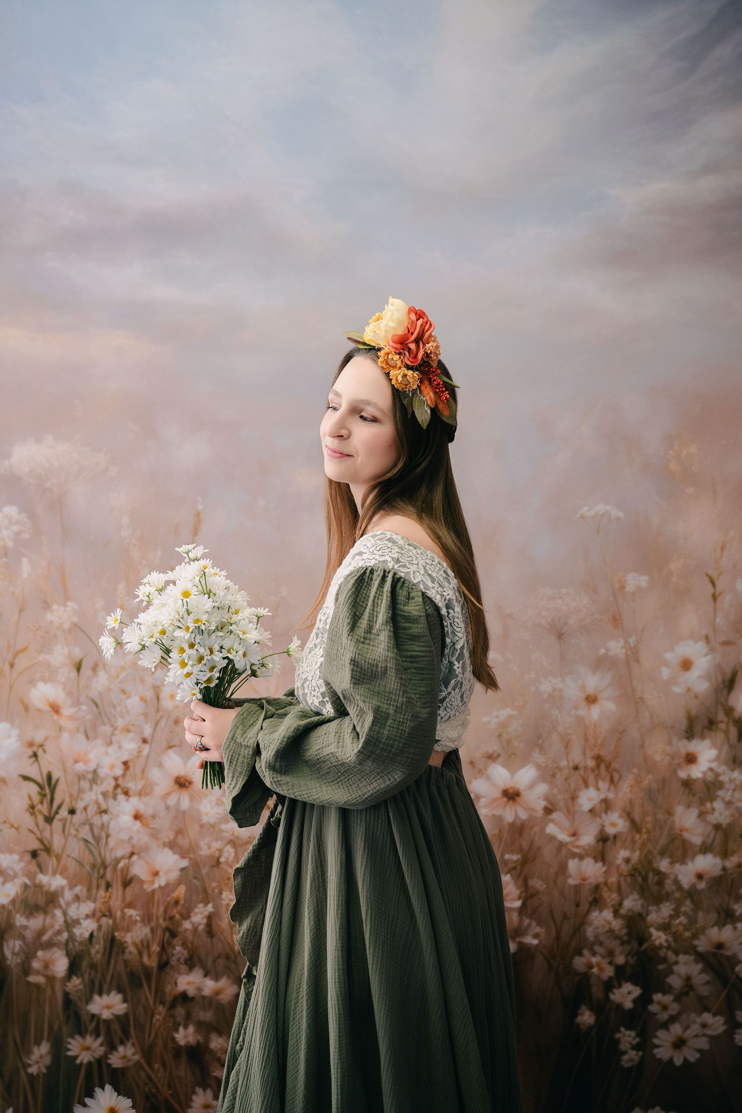 Young woman in green vintage dress holding white flowers, wearing floral headpiece, standing in dreamy meadow backdrop. mother day photoshoo