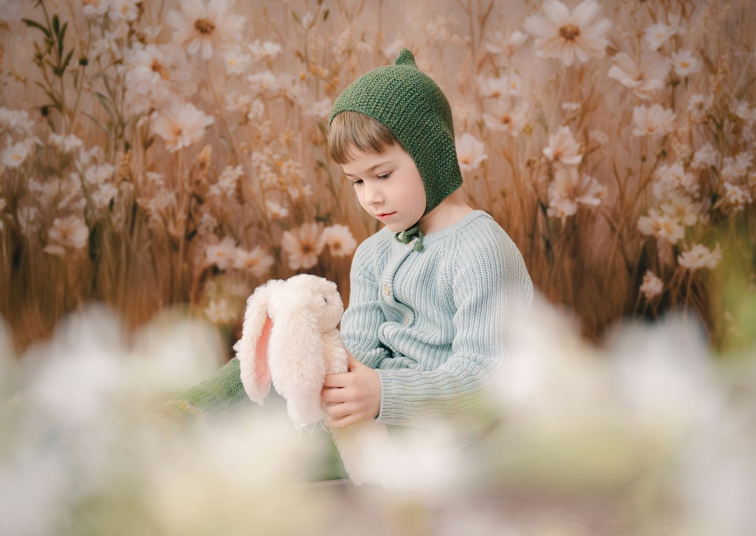 A child wearing a green knit hat sits among flower blossoms while holding a pink plush toy. Whimsical Portraits