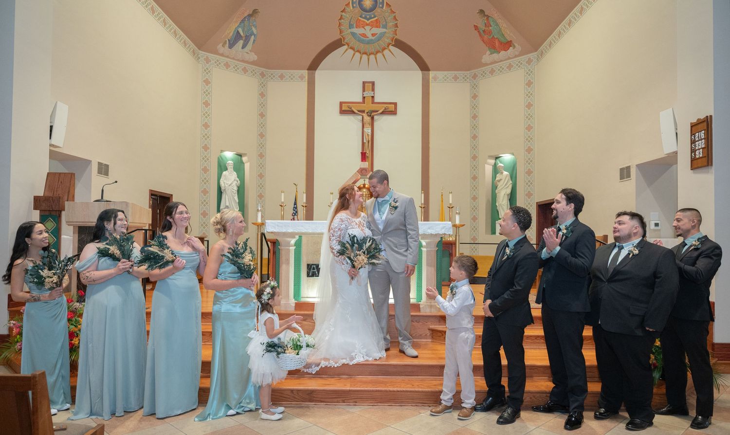Wedding party poses at church altar, bridesmaids in mint dresses, groomsmen in suits, bride and groom kissing.