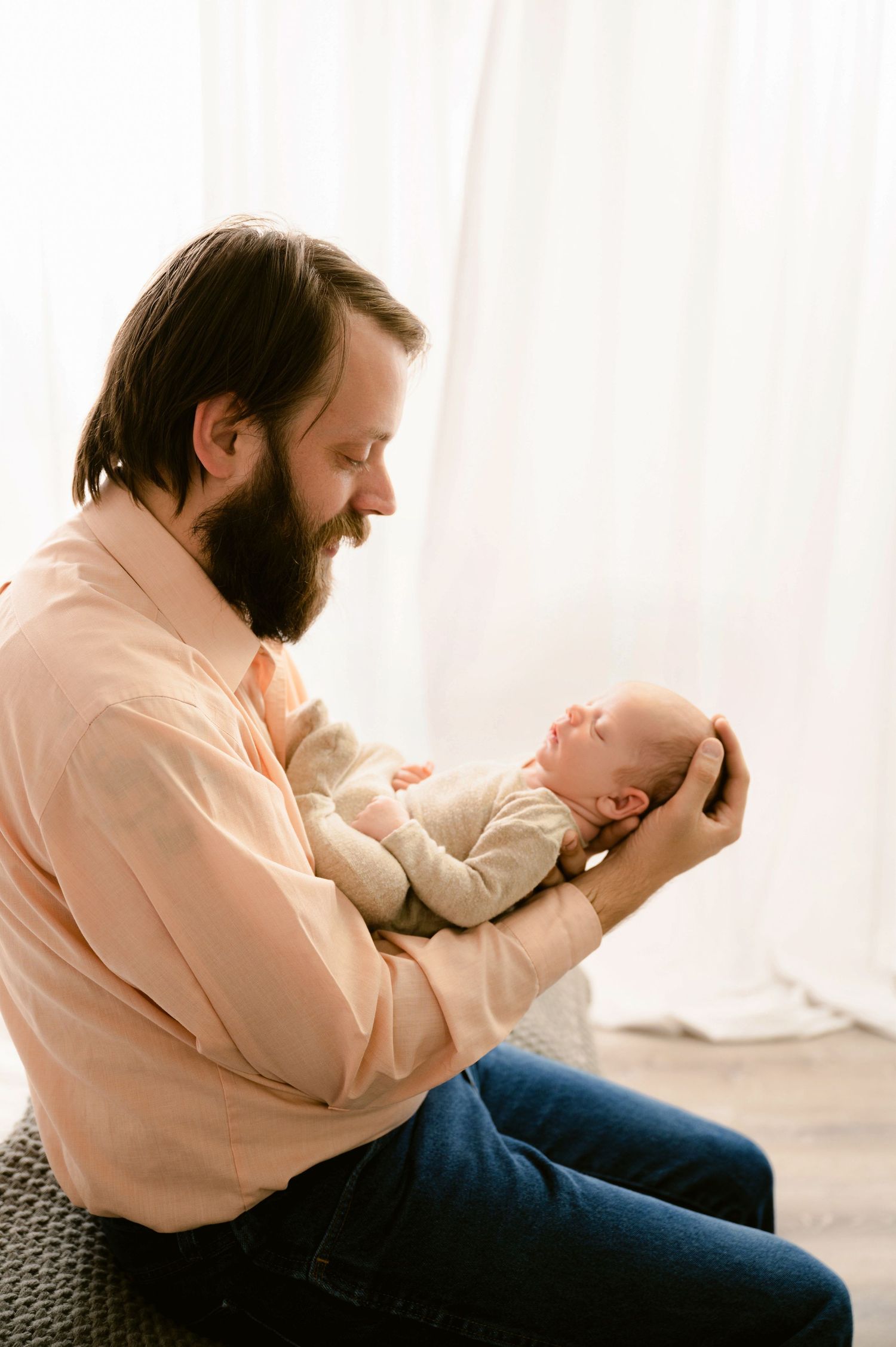A bearded man in a peach-colored shirt tenderly holds a newborn baby while sitting on a chair near a window.