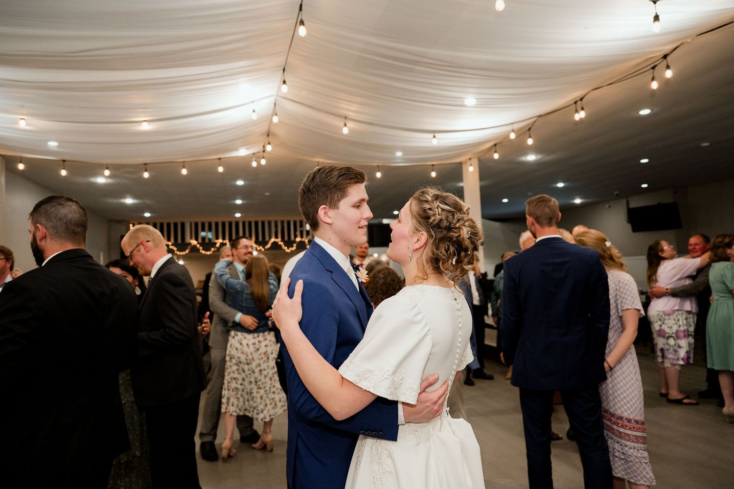 Wedding reception in a white draped venue with string lights and guests dancing on the dance floor.