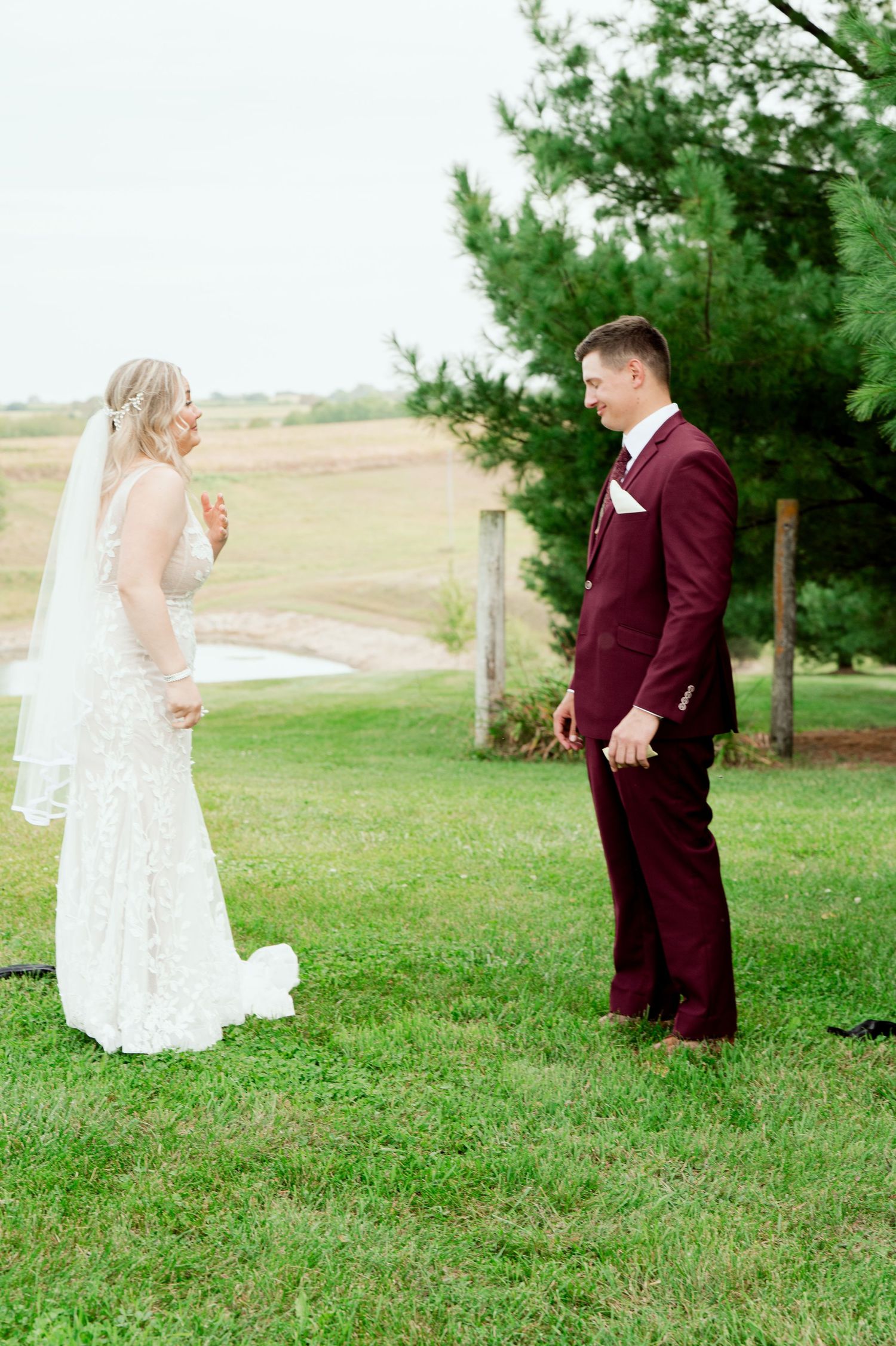 First look moment between bride and groom in burgundy suit on grassy hillside with evergreen trees.