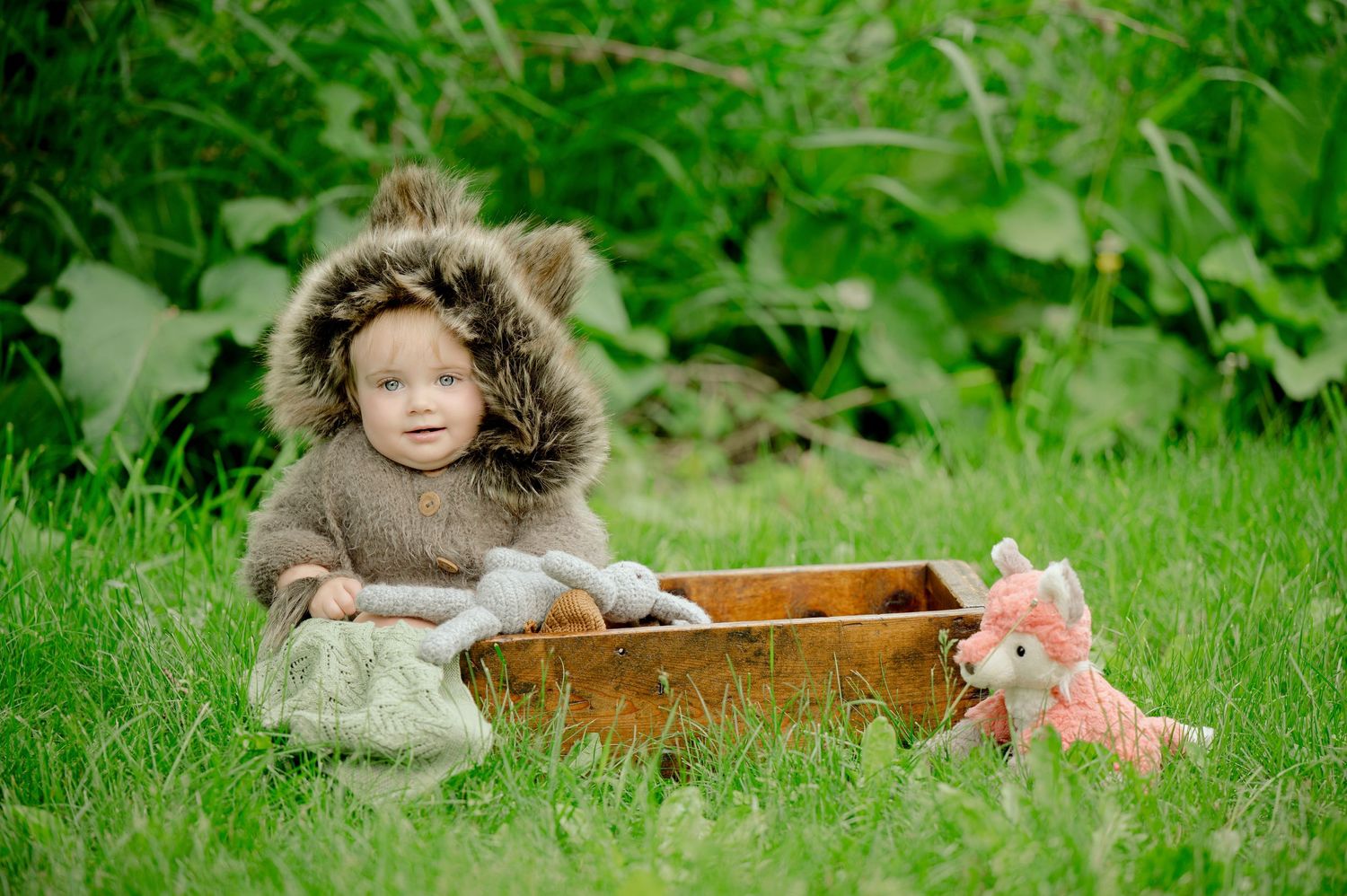 A small figure in a furry brown coat sits on grass beside a wooden box and pink plush toy in a garden setting.