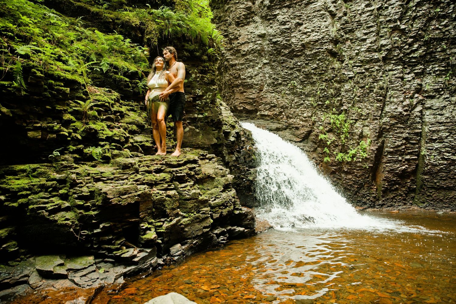 Couple embraces on mossy rocks beside a cascading waterfall in a lush green forest.