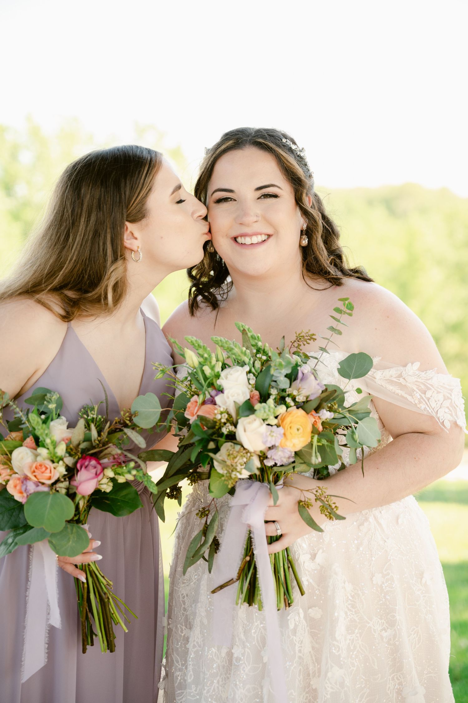 Central MN Wedding Photos - Bride in lace gown holds floral bouquet as bridesmaid in lavender dress kisses her cheek outdoors.