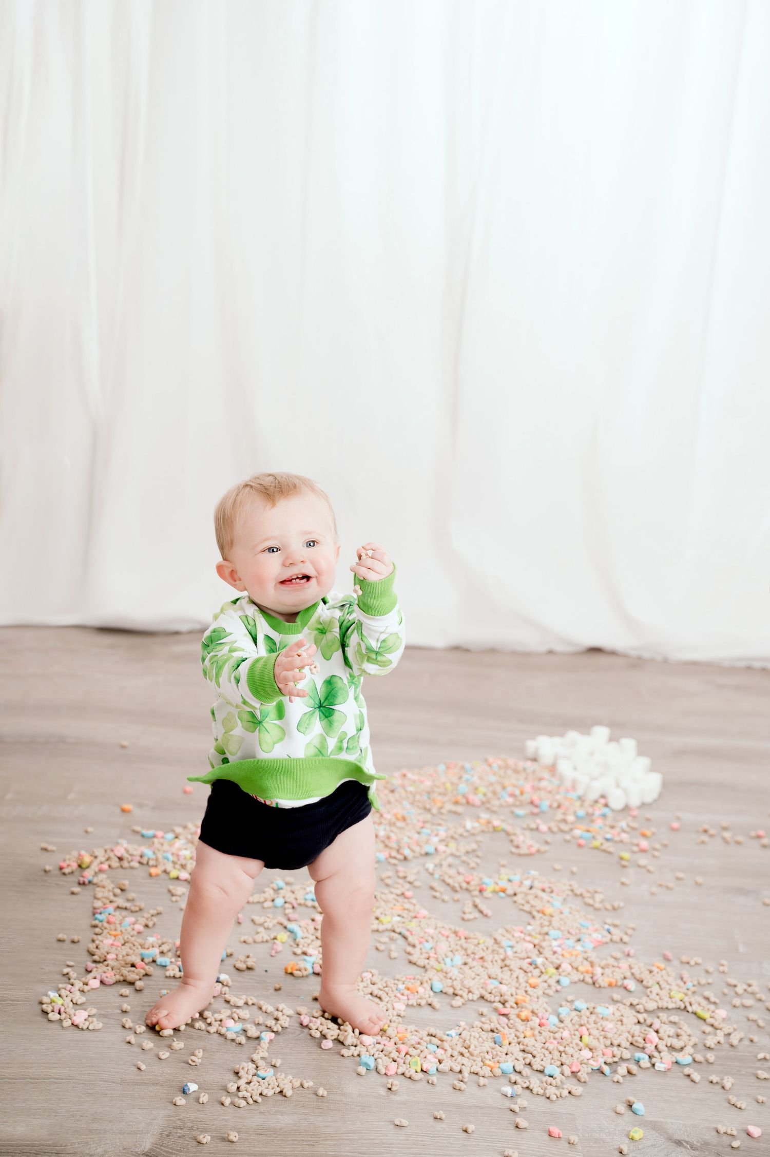 Central MN Milestone Photography - Baby in green outfit standing amid scattered colorful cereal on a white studio floor.