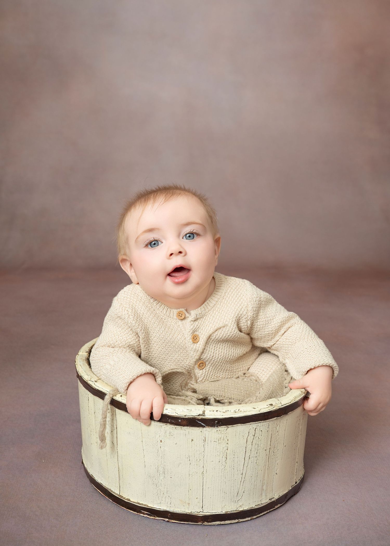 Central MN Milestone Photography - baby sits in a rustic white wooden bucket, wearing a cream knit outfit, against a soft brown backdrop.