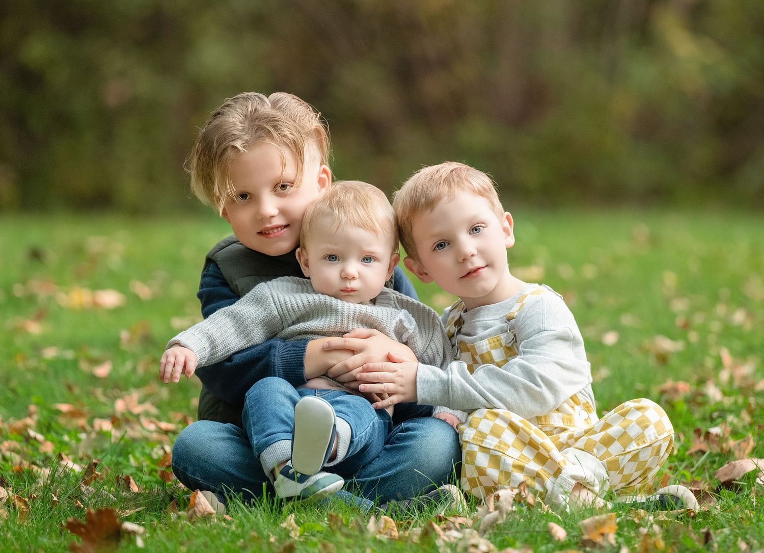 Three children sitting together on grass with autumn leaves in a garden setting.