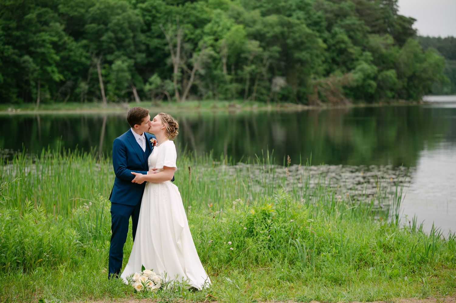 A wedding couple poses romantically by a tranquil lake surrounded by summer greenery.